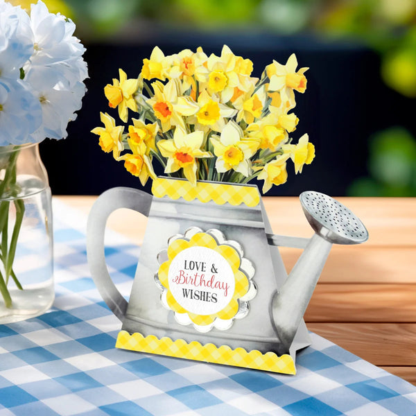 A decorative watering can-shaped card or gift holder filled with yellow daffodils, placed on a blue-and-white checkered tablecloth beside a glass vase of blue flowers. The front has a round label reading “Love & Birthday Wishes.