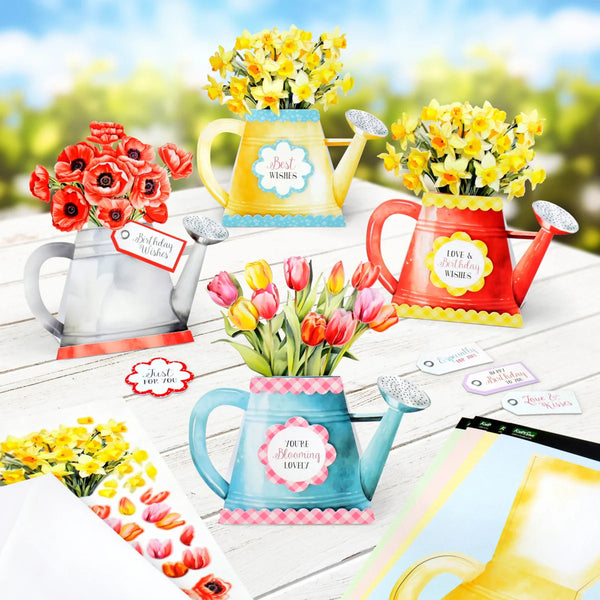 Colorful watering cans with flowers and decorative cards on a wooden surface.