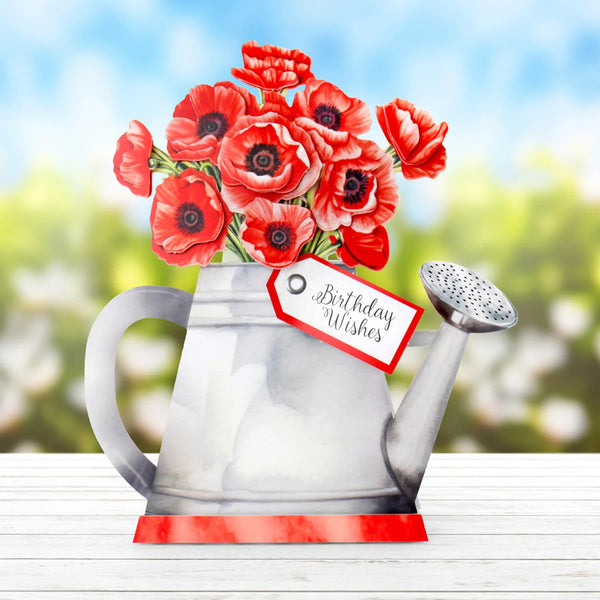 Red flowers in a watering can with a 'Birthday Wishes' tag against a blurred outdoor background