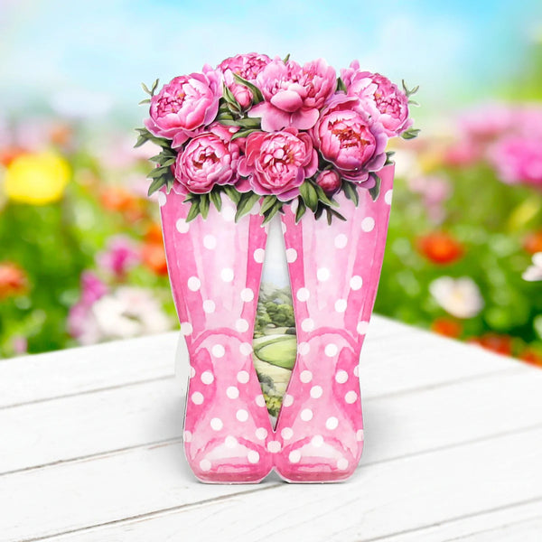 Pink polka dot rain boots with a bouquet of pink flowers on a white surface with a garden background.