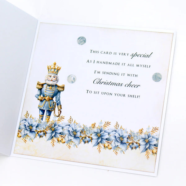An open white greeting card featuring a decorative nutcracker illustration in blue and gold, blue poinsettia flowers along the bottom, and printed holiday text on the front.