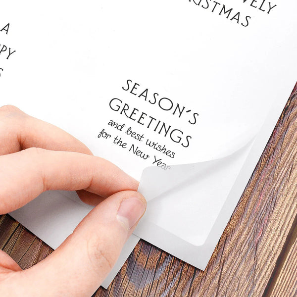 A close-up of a hand peeling back a transparent protective film from a white paper holiday greeting card placed on a wooden surface. The card shows printed text including 'SEASON'S GREETINGS' and partial 'MERRY CHRISTMAS'.