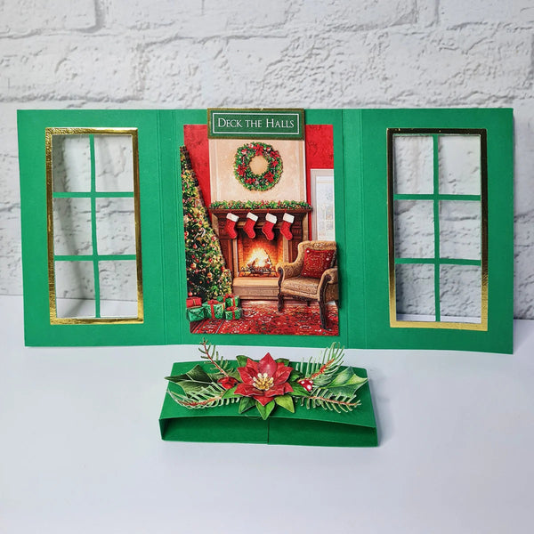 A green Christmas-themed card game set displayed on a white surface against a white brick wall. The set includes an open triptych-style card frame with gold-trimmed window cutouts and a central holiday scene, plus a matching green box with a red poinsettia and green leaf embellishment.