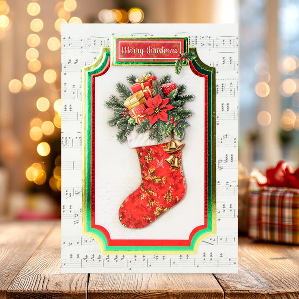 A festive Christmas greeting card displayed on a wooden surface, featuring a red patterned stocking filled with green pine branches, red poinsettias, and wrapped gifts, set against a sheet music background with warm bokeh lights.