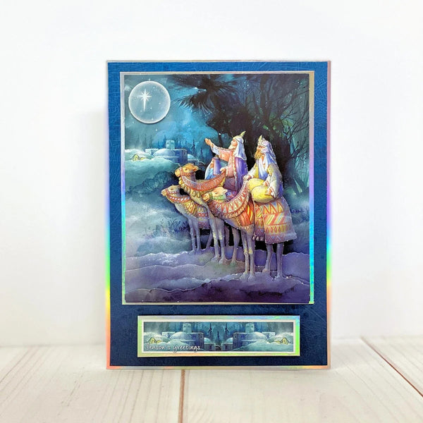 A vertical art print or card featuring the Three Wise Men under a full moon, with a holographic iridescent blue border and a small matching strip at the bottom, displayed on a light wood surface against a white wall.
