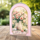 A decorative arch-shaped greeting card featuring a bouquet of pink roses in a vintage pitcher, displayed on a wooden surface with a blurred garden background. The card has a light pink border and the text 'Just for You' at the bottom.