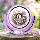 A layered circular card or decorative plaque with purple scalloped borders and a central floral wreath photo, displayed on a rustic wooden surface with blurred pink flowers in the background.