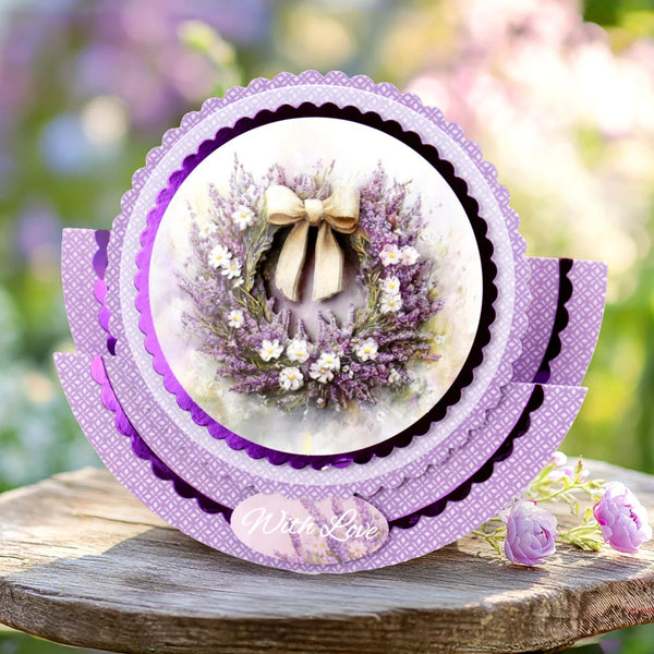 A layered circular card or decorative plaque with purple scalloped borders and a central floral wreath photo, displayed on a rustic wooden surface with blurred pink flowers in the background.