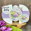 A handmade greeting card displayed on a rustic wood slice, featuring purple and lavender hydrangea illustrations, a purple polka dot pattern, and text reading 'Get Well Soon' and 'Love and Hugs'.