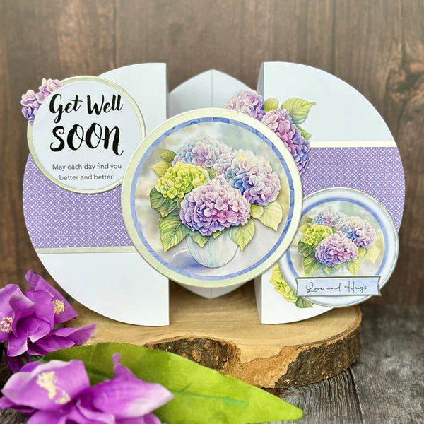 A handmade greeting card displayed on a rustic wood slice, featuring purple and lavender hydrangea illustrations, a purple polka dot pattern, and text reading 'Get Well Soon' and 'Love and Hugs'.