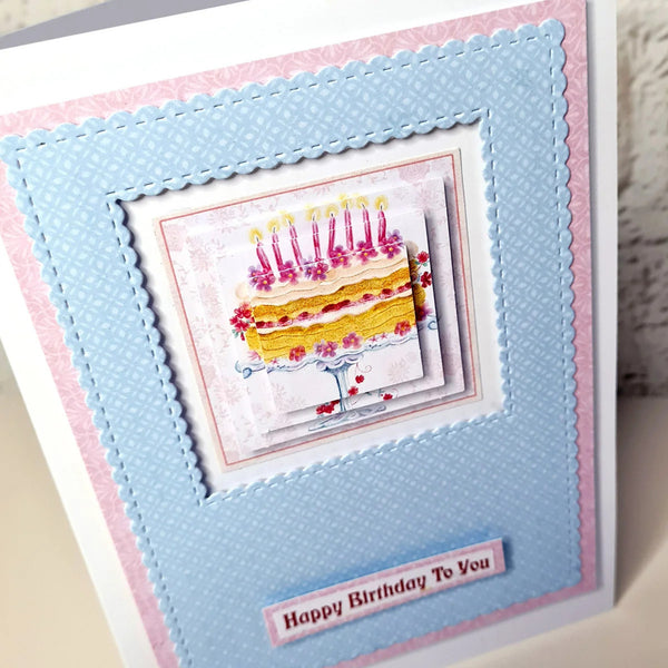 A handmade layered greeting card featuring a central illustration of a birthday cake with candles, surrounded by light blue and pink textured borders, with a small banner reading 'Happy Birthday To You'.