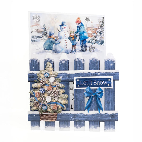 A decorative winter-themed card or art print featuring a snowy scene with children building a snowman, a blue picket fence, a small ornamented tree, and a blue bow with a sign reading 'Let it Snow'.