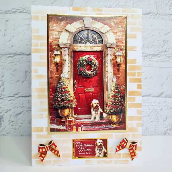 A handmade Christmas greeting card featuring a festive front door scene with a red door, wreath, decorated trees, and a small dog sitting at the door. The card is displayed on a white surface against a light gray brick-patterned background, with two red and gold ribbon bows at the bottom.