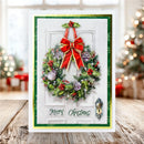 A Christmas greeting card displayed on a wooden surface with a blurred holiday background. The card features a white door design with a green wreath decorated with red berries, pinecones, and a large red bow, plus the text 'Merry Christmas' and a small ornament illustration.