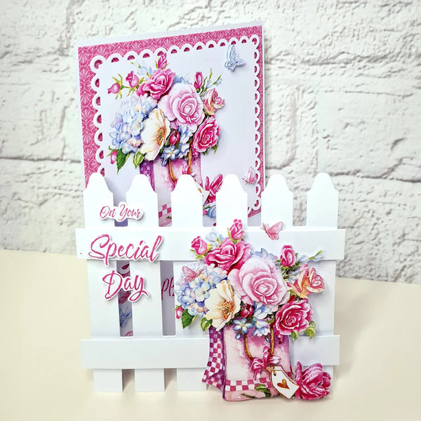 A handmade greeting card displayed on a white picket fence stand against a white brick wall. The card features a pink and white patterned border, a bouquet of pink and blue flowers, butterflies, and the text 'On Your Special Day'.