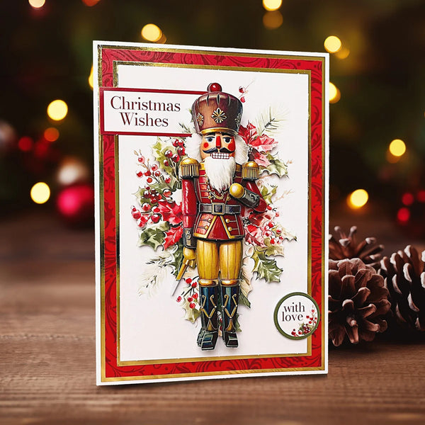 A handmade Christmas greeting card featuring a detailed nutcracker illustration with red and gold armor, surrounded by holly and red berries. The card has a red patterned border and the text 'Christmas Wishes' on the top left, standing on a wooden surface with pinecones and blurred holiday lights in the background.