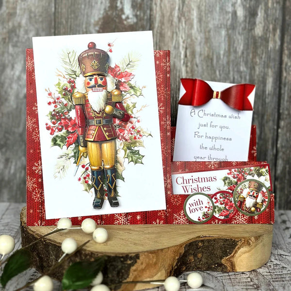 A Christmas greeting card set displayed on a rustic wood slice stand. The set includes a large card featuring a vintage nutcracker illustration with red berries and holly, a small red bow, a handwritten note, and a round sticker.