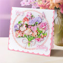 A square birthday card with a pink and white scalloped border, featuring a 3D layered bouquet of pink and purple flowers with green stems, a butterfly, and the words 'Happy Birthday' in pink script. The card is displayed on a light surface next to a glass vase with flowers.