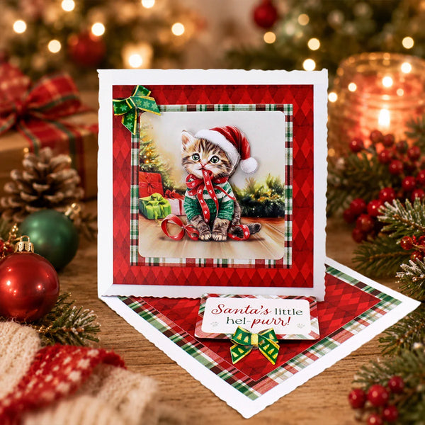 A square Christmas greeting card with a kitten in a Santa hat, displayed on a wooden surface with holiday decorations including ornaments, pine branches, and a candle in the background.