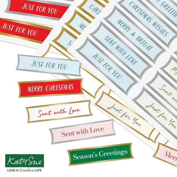 A collage of decorative holiday label stickers with phrases like 'Just for You,' 'Merry Christmas,' 'Sent with Love,' and 'Season's Greetings' in red, blue, pink, green, gold, and gray on a white background. The Katy Sue brand logo appears in the lower left corner.