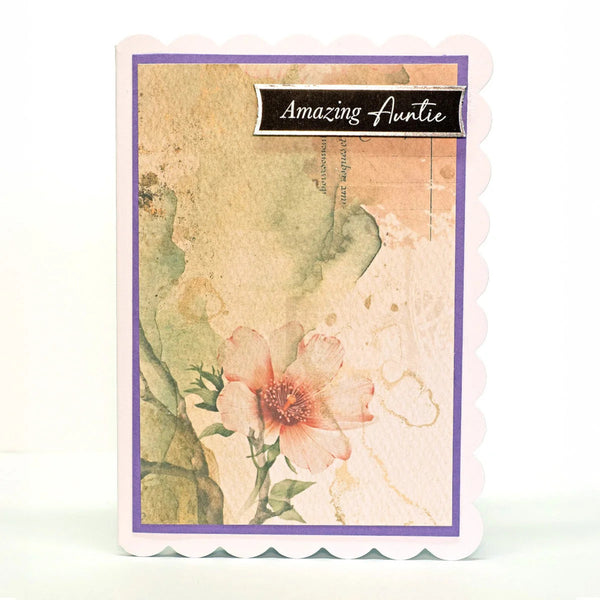 A handmade greeting card with a scalloped white border, featuring a vintage-style floral design with a pink flower, green leaves, and a purple inner border. A black banner at the top reads 'Amazing Auntie'.