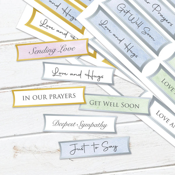 A set of printed sympathy and support message labels or stickers arranged on a white wood surface. The labels have soft pastel colors including pink, gold, blue, green, and gray, with handwritten-style text such as 'Love and Hugs,' 'Sending Love,' 'In Our Prayers,' 'Get Well Soon,' 'Deepest Sympathy,' and 'Just to Say.