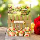 A handmade-style birthday card featuring a brown picket fence, potted roses, and colorful butterflies, displayed on a wooden surface with red and yellow roses in the foreground.