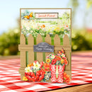 A handmade birthday card featuring a brown picket fence, colorful flowers, a red watering can, and butterflies, displayed on a red and white checkered tablecloth with a blurred garden background.