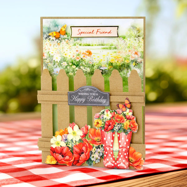 A handmade birthday card featuring a brown picket fence, colorful flowers, a red watering can, and butterflies, displayed on a red and white checkered tablecloth with a blurred garden background.