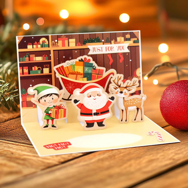A 3D pop-up Christmas greeting card displayed on a wooden table, featuring cartoon characters including Santa Claus, an elf, and a deer in front of a festive gift shop background with warm string lights.
