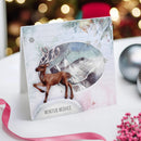 A handmade Christmas greeting card featuring a brown reindeer standing in front of a snowy mountain scene, displayed on a white surface with festive blurred lights in the background.