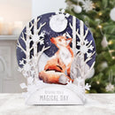 A decorative tabletop holiday display featuring a watercolor fox in a snowy winter scene with birch trees, snowflakes, and a moon, plus a white base that reads 'Wishing you a Magical Day.' The background shows a softly blurred Christmas tree and indoor setting.
