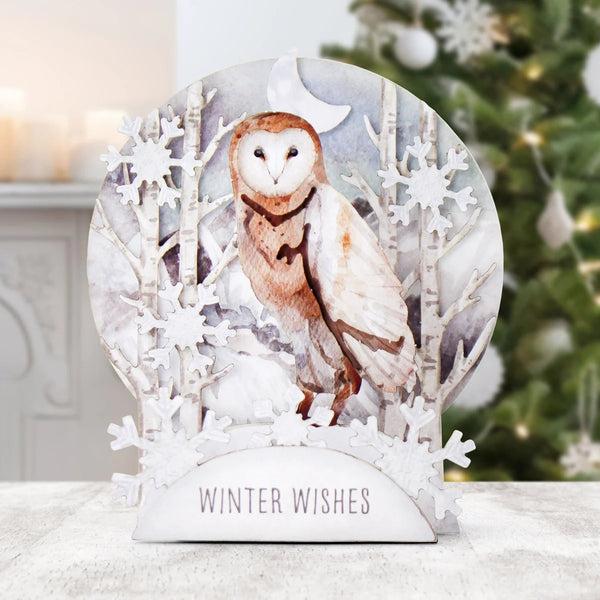 A decorative winter-themed tabletop sign featuring a painted barn owl in a snowy forest scene with snowflakes and birch trees, mounted on a white base that reads “WINTER WISHES.” The sign is photographed on a light surface with a softly blurred Christmas tree and indoor background.
