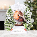 A Christmas-themed tabletop decoration featuring a watercolor robin perched on a log between two snowy pine trees, with white snowflakes, holly leaves, and a banner reading “MERRY CHRISTMAS.” The background shows a softly blurred indoor holiday scene with a Christmas tree and warm lights.