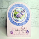 A handmade sympathy card with an arched top and a pink patterned base. The center features a circular layered design with blue paper flowers, a purple hydrangea, green hydrangea, and a butterfly.