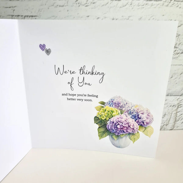 A white greeting card shown open against a white brick wall background. The card features the text 'We're thinking of You and hope you're feeling better very soon.