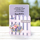 A handmade birthday greeting card with a pink picket fence design, floral bouquet, and bird illustration, displayed on a white wooden surface with a blurred green outdoor background.