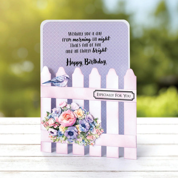 A handmade birthday greeting card with a pink picket fence design, floral bouquet, and bird illustration, displayed on a white wooden surface with a blurred green outdoor background.