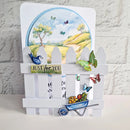 A handmade garden-themed greeting card featuring a white picket fence, a blue bird, colorful butterflies, a small wheelbarrow, and a circular landscape illustration in the background.
