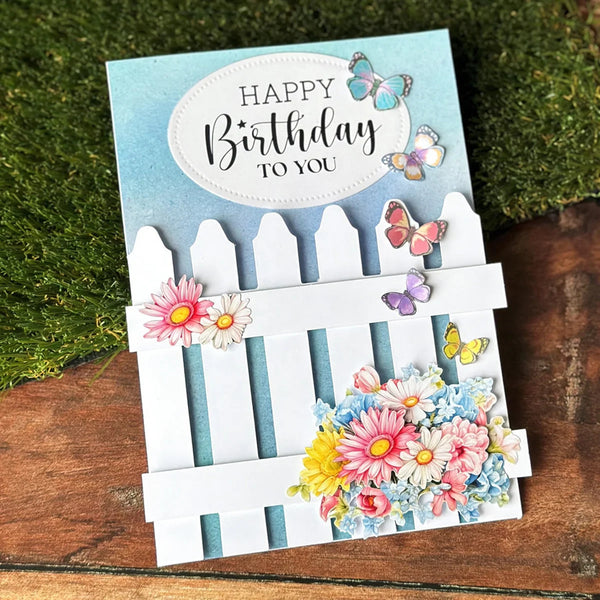 Birthday card with flowers, butterflies, and a white picket fence design on a wooden surface.