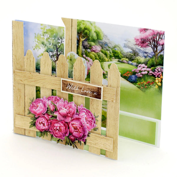 A handmade greeting card or art print featuring a light wood fence in front of a colorful garden scene with pink peonies. The front of the fence has a small wooden sign reading 'With Love, x'.