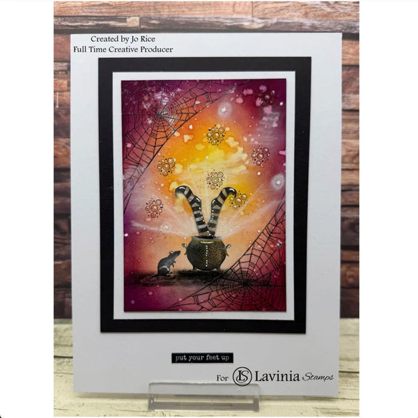 A handmade greeting card featuring a whimsical Halloween design with a cat inside a cauldron, spiderwebs, and a glowing orange and purple sky. The card is displayed on a small clear stand against a rustic wood plank background.