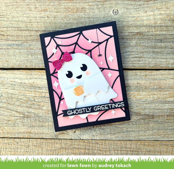 A handmade Halloween greeting card featuring a cute white ghost holding a pumpkin on a pink spiderweb background, placed on a rustic wooden surface. The card has a black border and a black banner that reads 'GHOSTLY GREETINGS'.