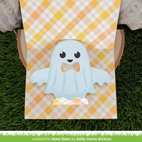 A cute ghost-shaped decoration or card featuring a light blue ghost with a yellow bow tie, displayed on a yellow and white gingham background with a green grass-like surface.