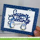 A handmade greeting card held in a hand, featuring the words 'Seasons Greetings' in blue script over a white textured background, framed by a navy blue border with white polka dots. Two small blue and white penguin illustrations are at the bottom, with white snowflake accents.