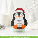 A handmade Christmas penguin card gift tag with a red Santa hat, displayed on a small clear stand. The card features a black and white penguin design with orange feet and beak, and the text 'Merry Wishes To FROM'.