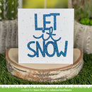 A square white card with glittery blue text reading “LET it SNOW” and small snowflake details, displayed on a round wood slice over artificial grass with blurred potted plants in the background.