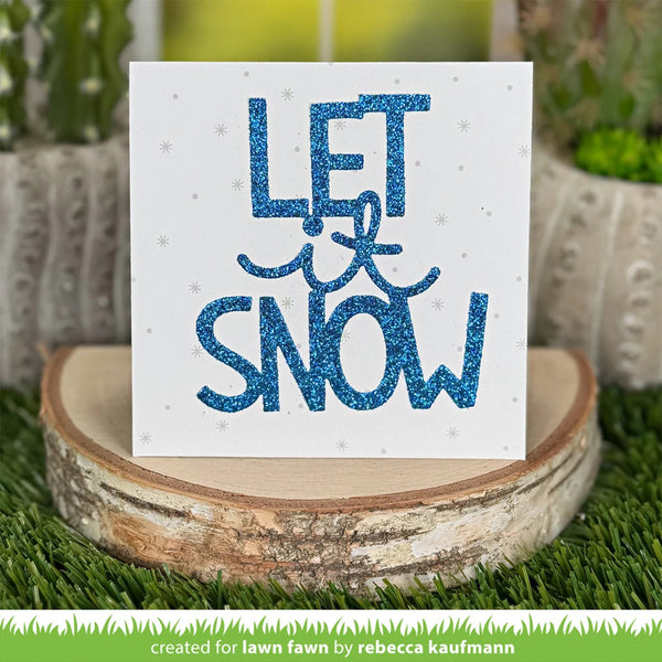 A square white card with glittery blue text reading “LET it SNOW” and small snowflake details, displayed on a round wood slice over artificial grass with blurred potted plants in the background.