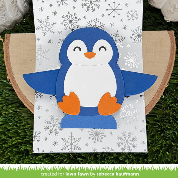 A handmade penguin card or decoration made from layered blue, white, and orange paper, displayed on a patterned white sheet over a wood slice and artificial green grass. A green banner at the bottom reads 'created for lawn fawn by rebecca kaufmann'.