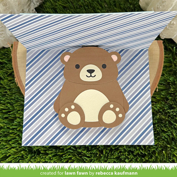 A handmade paper pop-up gift box featuring a cute brown bear with a cream belly and black nose, set against a blue and white striped background. The box is displayed open on a green grass-like surface.